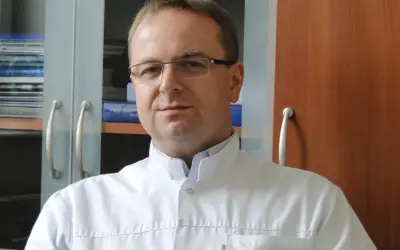 Portret mężczyzny w okularach i białym fartuchu lekarskim, na tle regałów z książkami.