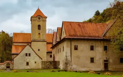 Czerwony klasztor w Pieninach, z charakterystyczną wieżą zegarową i pomarańczowymi dachami, otoczony zielonym trawnikiem i drzewami.