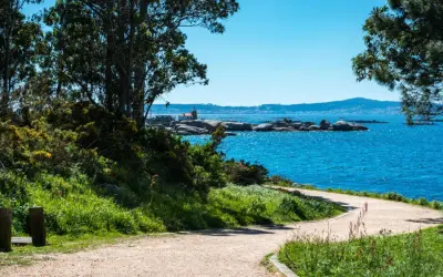 Sendero costero en la Illa de Arousa con mar azul brillante, rocas y un pequeño faro.
