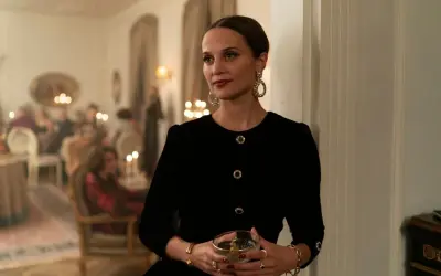 Alicia Vikander i en elegant svart klänning med stora örhängen, håller ett glas. Scenen utspelar sig på en tillställning, kanske från en av hennes filmer.