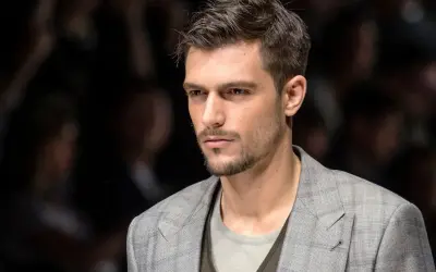 Uomo con capelli spettinati corti, sguardo intenso, indossa una giacca a quadri grigia.