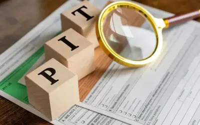 Drewniane klocki z literami "PIT" leżą na formularzu podatkowym. Lupa bada szczegóły.