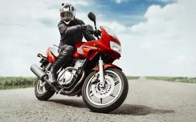 Motociclista con casco e giacca in pelle, in posizione di guida sportiva su una moto rossa su una strada vuota.