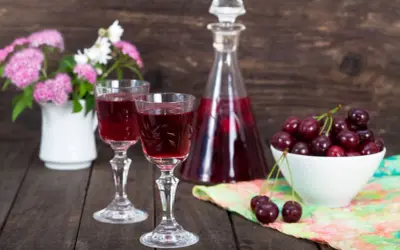 Dwa kieliszki z rubinową nalewką z czereśni na bimbrze, obok miseczka świeżych owoców i bukiecik kwiatów.