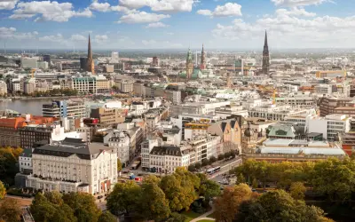 Blick von einer **Aussichtsplattform Hamburg** über die Stadt mit ihren Kirchtürmen, dem Hafen und grünen Bäumen im Vordergrund.