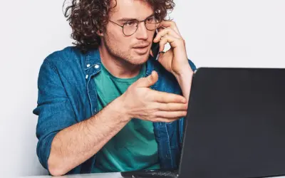 Mężczyzna w okularach rozmawia przez telefon, gestykulując w stronę laptopa. Szuka uczciwych firm oddłużeniowych, by rozwiązać swoje problemy finansowe.