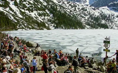 Tłum ludzi odpoczywa nad zamarzniętym jeziorem Morskie Oko, w tle góry pokryte śniegiem i lasem. Palenica Białczańska w oddali.