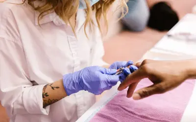 Manicurzystka w niebieskich rękawiczkach precyzyjnie usuwa skórki wokół paznokci klienta za pomocą cążków.