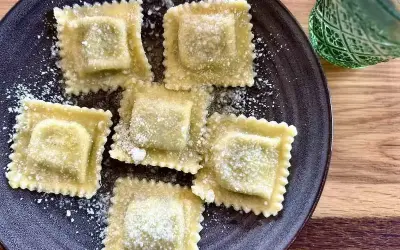 Pięć kwadratowych ravioli z ricottą, posypanych parmezanem, na ciemnym talerzu. Obok zielony kieliszek.
