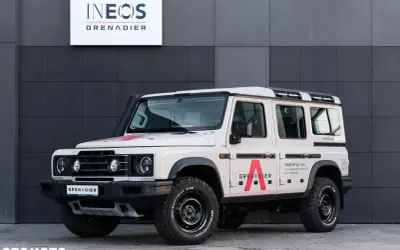Beżowy INEOS Grenadier z czerwonym logo "A" na drzwiach. Sprawdź INEOS Grenadier cena i możliwości tego terenowego SUV-a.