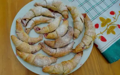 Pyszne, domowe rogaliki z marmoladą, posypane cukrem pudrem, ułożone na talerzu obok ręcznika z truskawkami.
