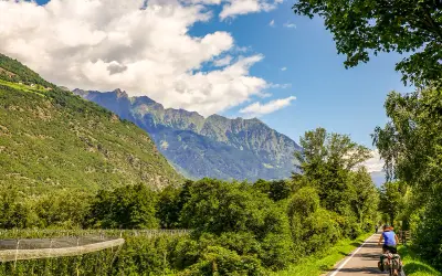 Cycliste sur la Via Claudia Augusta, traversant une vallée verdoyante bordée de montagnes majestueuses.