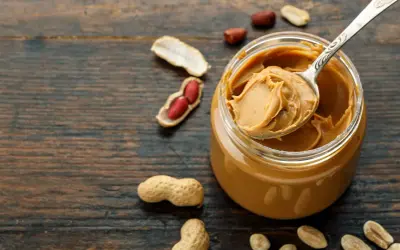 Ein Löffel cremiger Erdnussbutter aus einem Glas, umgeben von Erdnüssen. Erdnussbutter gesund und lecker!