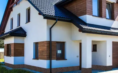 Nowy dom z białym tynkiem i drewnianymi akcentami. Brak cokołu na elewacji, widoczna kamyczkowa podmurówka.