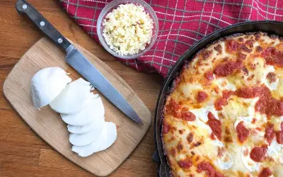 Idealny jaki ser do pizzy? Na desce świeża mozzarella, obok nóż i miseczka z tartym serem. Obok pyszna, gorąca pizza.
