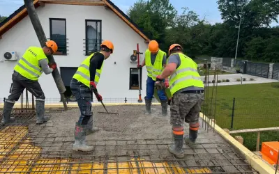 Budowlańcy w kamizelkach i kaskach, przestrzegając zasady bhp, wylewają beton. Jeden kieruje rurę, drugi grabi, trzeci wyrównuje.