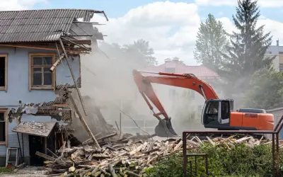 Pomarańczowy koparka prowadzi rozbiórkę budynku, wzbijając tumany kurzu. Ruiny i drewno leżą na ziemi.