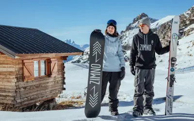 Para trzymająca deski snowboardowe przed drewnianą chatką w górach.