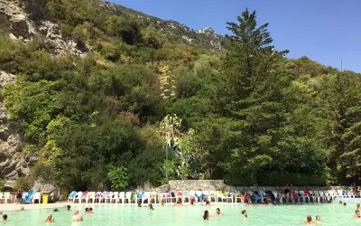 Piscina con persone che nuotano, circondata da vegetazione lussureggiante e rocce, un luogo che sembra uscito dalle grotte delle ninfe.
