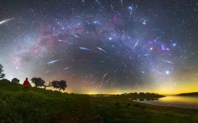 A lone figure watches a spectacular display of shoot stars streaking across the Milky Way above a tranquil lake and grassy hills.