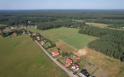 Widok z lotu ptaka na wieś otoczoną lasem. Domy z czerwonymi dachami i zielone pola. Teren ma około 1 ar.