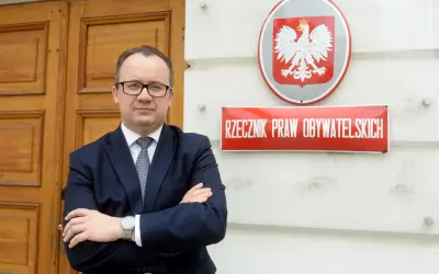 Mężczyzna w garniturze stoi przed budynkiem z godłem Polski i tabliczką "Rzecznik Praw Obywatelskich".