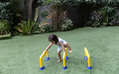 A toddler practices gross motor outdoor activities, balancing on yellow and blue hurdles in a grassy yard.