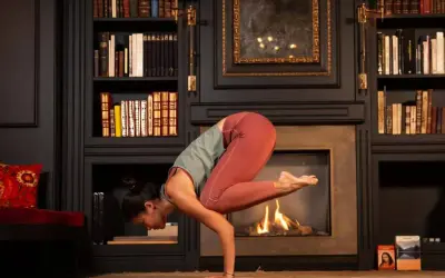Mujer en postura de grulla, inspirada en frases yoga Patanjali. Chimenea encendida y estanterías llenas de libros.