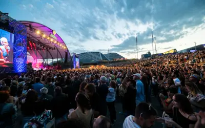 Multitud disfrutando de un concierto al aire libre. Se ve un gran escenario con luces y pantallas, y miles de personas animando. ¡Así se vive latino Zaragoza!