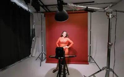 Modelka w pomarańczowej sukience pozuje na tle czerwonego tła. Wokół niej profesjonalne **światło studyjne** i sprzęt fotograficzny.