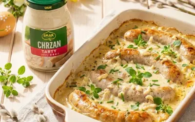 Biała kiełbasa w sosie chrzanowym, zapiekana w naczyniu, z dodatkiem ziół. Obok słoik sosu.