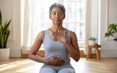 Femme pratiquant une technique de relaxation mentale, mains sur la poitrine et le ventre, yeux fermés, dans une pièce lumineuse.