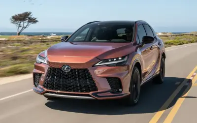 Miedziany Lexus RX 500h jedzie wzdłuż wybrzeża. Słońce odbija się od jego lśniącej karoserii.