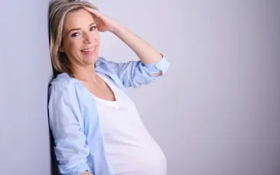 Uśmiechnięta kobieta w ciąży po 40. opiera się o ścianę, dotykając włosów.