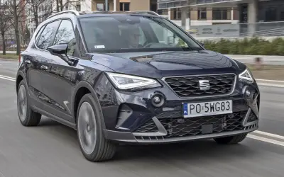 Ciemnoniebieski Seat Arona w ruchu ulicznym. Kierowca w okularach. Wiele pozytywnych seat arona opinie potwierdza jego miejski charakter.