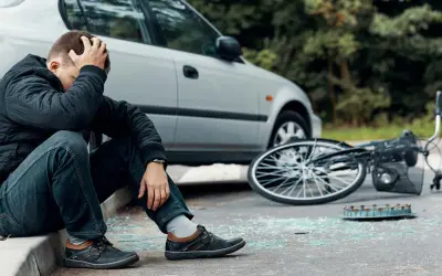 Man zit op stoeprand, hand in haar, na een fietser aangereden te zijn. Fiets ligt omgevallen, met glasscherven op straat.