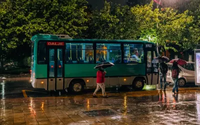Deszczowa noc w Tiranie. Autobus z napisem "Dalje" i "Hyrje" stoi na mokrej ulicy, ludzie z parasolami czekają na przystanku.