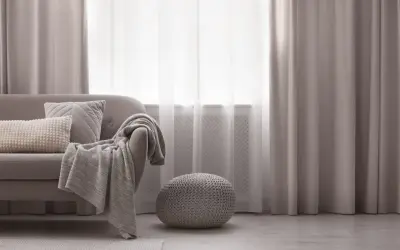 Elegancka dekoracja okien w salonie: szare zasłony i firanki tworzą przytulną atmosferę. Sofa z poduszkami i pledem zaprasza do relaksu.