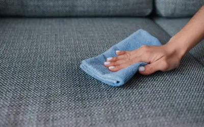 A hand with white nail polish wipes a grey couch with a blue microfiber cloth, demonstrating how to remove slime from couch.
