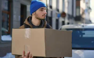 Młody mężczyzna w niebieskiej czapce niesie paczkę. Czy dpd dostarcza w soboty? Zima na ulicy, śnieg na chodniku.