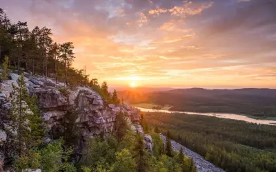 Solnedgång över en skogklädd dal med en sjö. Klippor och träd i förgrunden. En vacker vy som ger fakta om Finland.