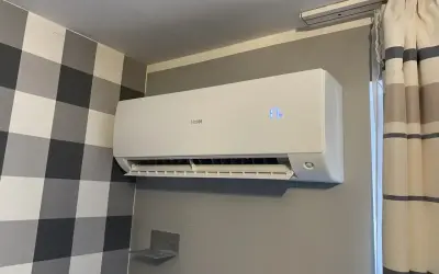 Biała klimatyzacja Haier zamontowana na szarej ścianie, obok zasłony.