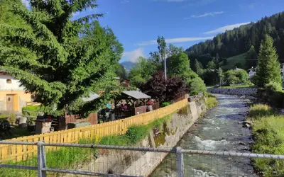 Un ruscello scorre a Santo Stefano di Cadore, fiancheggiato da case, alberi e un'area con gazebo e tavoli all'aperto.