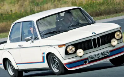 Białe BMW 2002 Turbo z kolorowymi pasami na zderzaku i błotnikach, pędzi po torze.