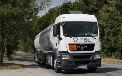 Biały cysterna z logo TSL jedzie drogą wśród drzew. To ważny element branży TSL, transportującej towary.