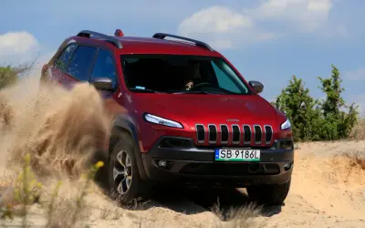 Czerwony Jeep Cherokee Trailhawk z impetem przemierza piaszczysty teren, wzbijając tumany piasku.