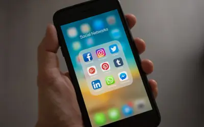 Dłoń trzyma smartfon z ikonami aplikacji społecznościowych. Widoczna ikona Facebooka, która pomoże Ci dowiedzieć się, jak dołączyć do grupy na FB.