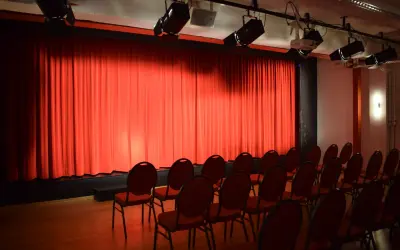 Rote Vorhänge und leere Stühle im **theas theater**. Scheinwerfer sind an der Decke montiert, bereit für die nächste Vorstellung.
