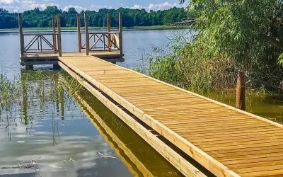 Drewniany pomost nad jeziorem zaprasza do spaceru. Woda odbija błękitne niebo i zielone drzewa.