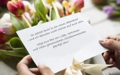 Hände halten eine Karte mit Hochzeitsglückwünschen vor Tulpen. "Ihr startet heute in ein neues Abenteuer – und wir könnten nicht stolzer auf euch sein. Möge eure Ehe von Liebe, Vertrauen und vielen gemeinsamen Glücksmomenten geprägt sein!